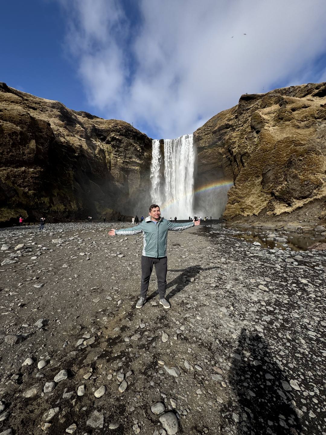 A traveler standing with arms wide in front of Skógafoss as a rainbow forms in the spray